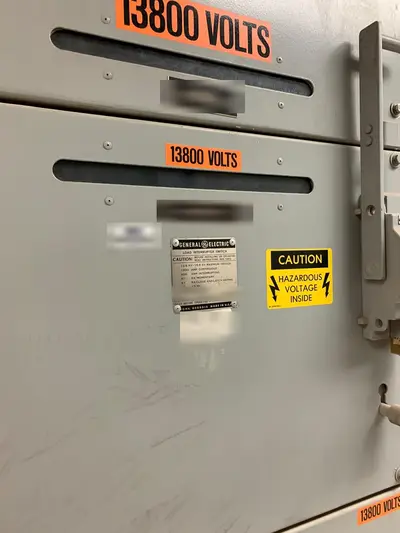 Switchgear cubicle doors marked 13800 VOLTS above and below the seam, with the General Electric load-interrupter-switch rating plate and a hazardous-voltage warning between them. Equipment designation and asset plates are masked.