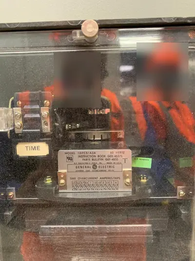 A General Electric inverse-time overcurrent relay behind its glass cover, tap block and time dial on the face, model and instruction-book data printed on the plate below. Reflections in the glass are masked.
