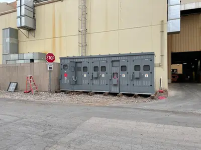 An outdoor metal-enclosed switchgear lineup set on timbers against a mill wall, four sections with viewing windows, arc-flash labels and operating handles, one door standing open.
