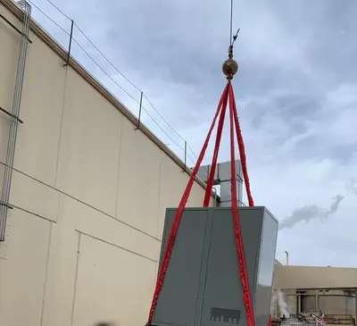 A switchgear section slung on four synthetic round slings from a crane hook, lifted clear of the ground alongside the building it is being set into.