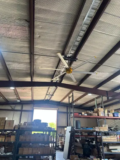 A high-volume low-speed fan and a run of high-bay strip lighting hung from the purlins of an occupied warehouse, the feeds carried along the beam line. A sign above the roll-up door is masked.