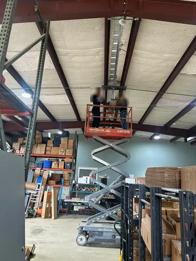 Two electricians working from a scissor lift at deck level above a stocked aisle, making off the drop for an overhead fan. Faces are masked.