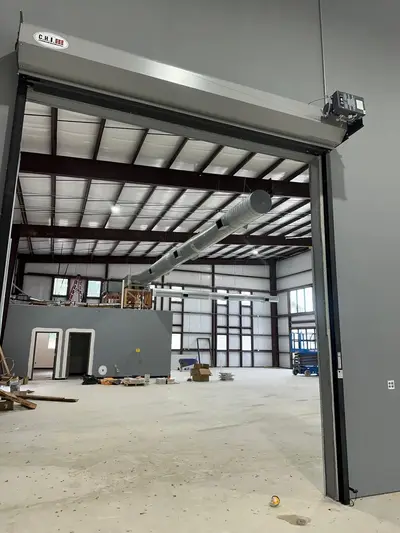 A metal building at fit-out stage: sectional overhead door in place, spiral duct hung, and the lighting circuits being run from a lift.