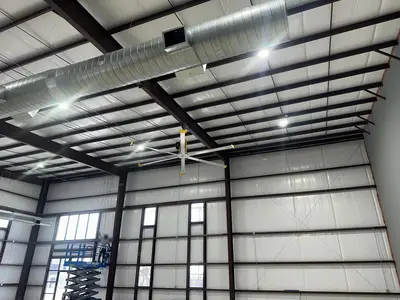 High-bay fixtures and a high-volume low-speed fan hung between the roof purlins of a new metal building, the main duct run crossing above. The worker on the lift is masked.