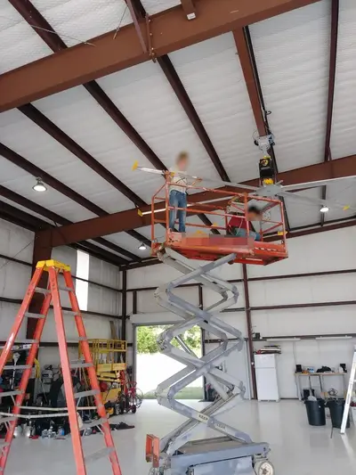 Assembling a high-volume low-speed fan from a scissor lift under the ridge of a hangar, blades fitted to the hub one at a time. Faces are masked.