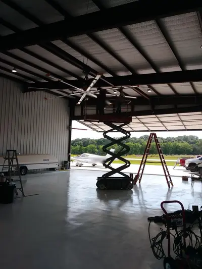 A completed high-volume low-speed fan on its ridge mount inside a hangar, the lift still standing beneath it and the apron visible through the open door. Faces and an aircraft registration are masked.