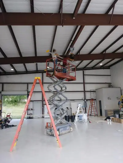 Two electricians at height on a scissor lift, working a fan mount into the ridge steel of the hangar. Faces are masked.