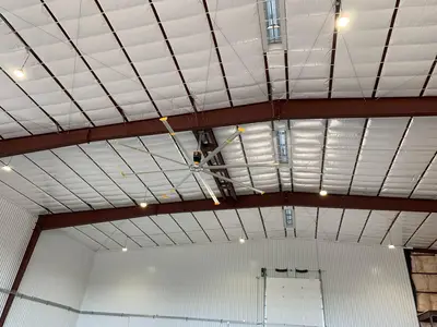 Looking up into the hangar roof at the fan on its ridge mount and the high-bay fixtures hung between the frames.