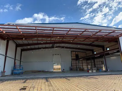 The hangar seen across the reinforced slab before the pour, roof framing and lighting complete inside the open bay. A figure inside the bay is masked.